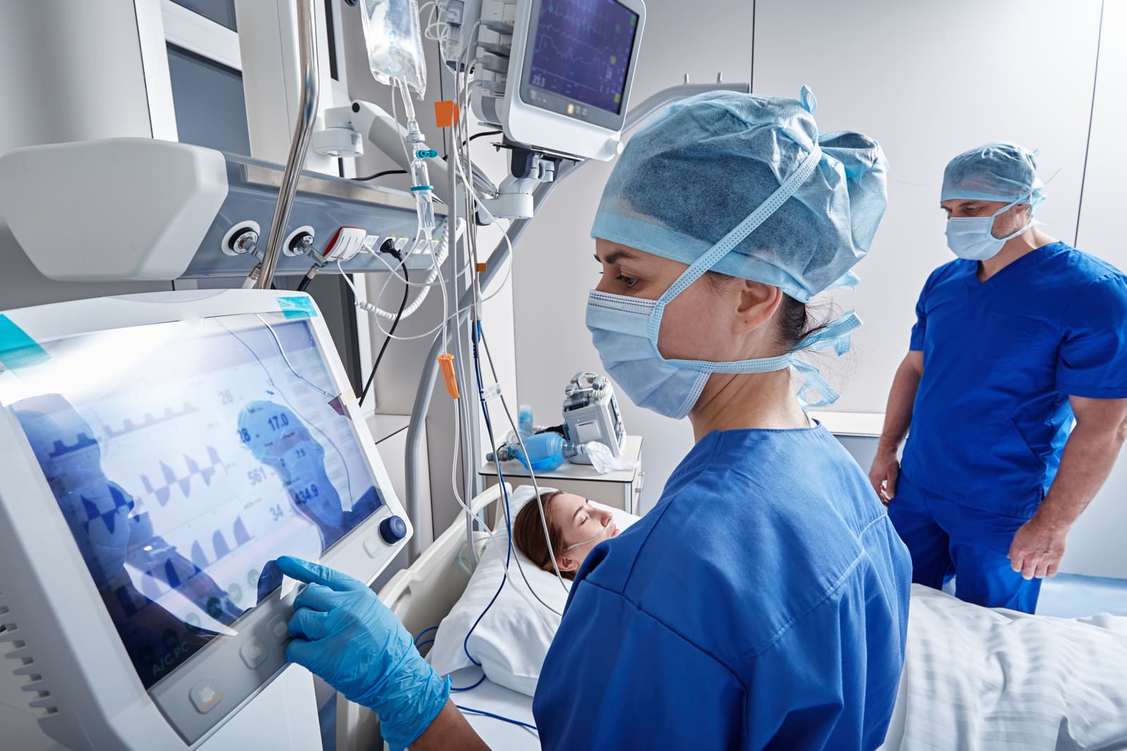 A clinician in a cap and mask using a touchscreen ultrasound system beside a patient