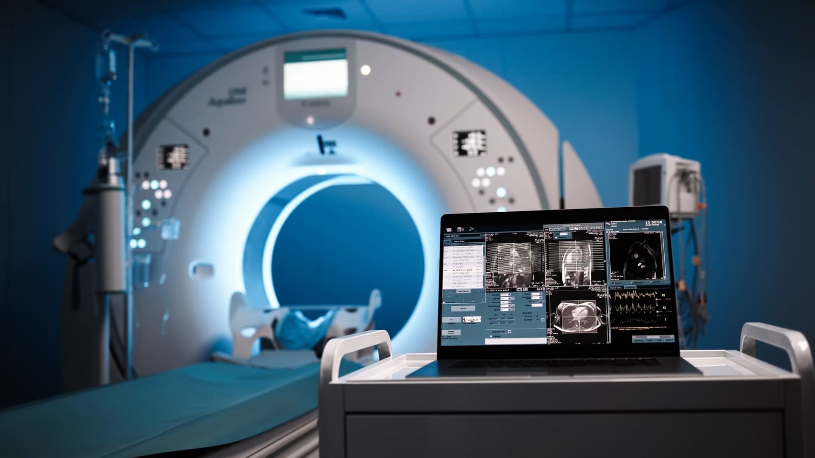 An MRI scanner in a bright imaging suite, with a laptop showing scan images in the foreground
